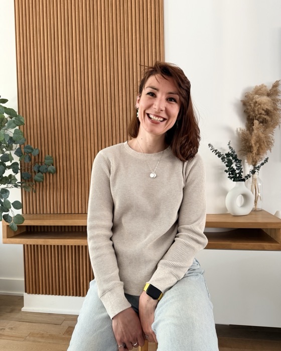 Pascaline assise sur un tabouret en bois, souriante, devant un mur de panneaux de bois et un eucalyptus