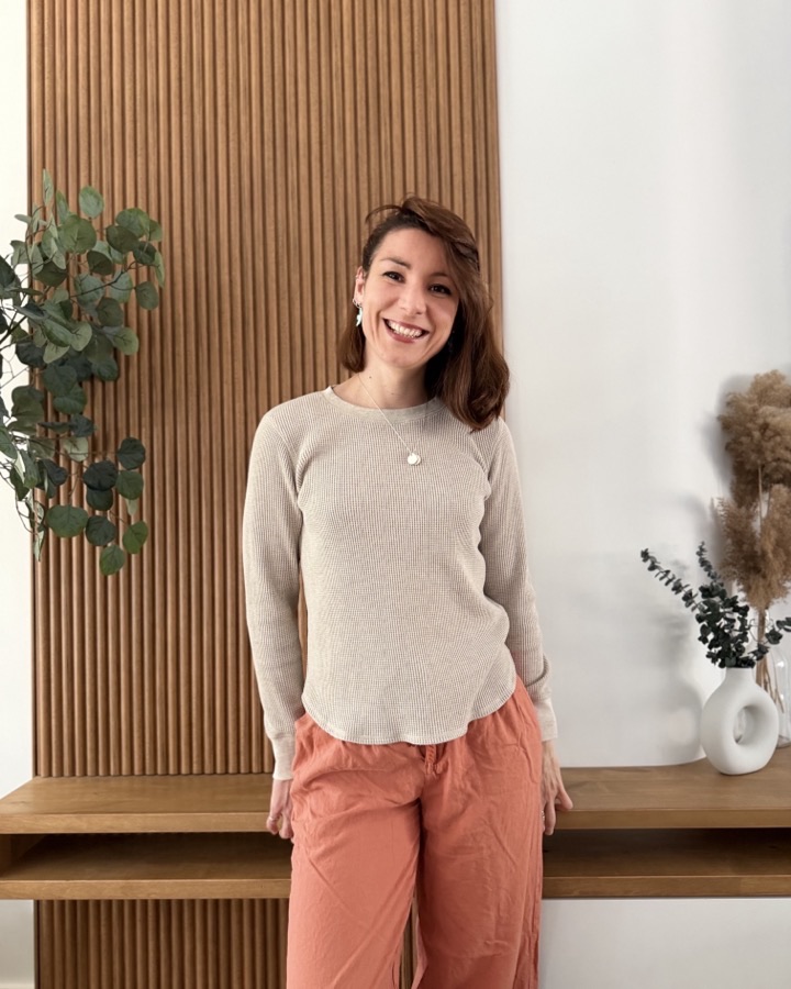 Pascaline souriante, debout devant un mur de bois clair avec un eucalyptus et un vase de pampas